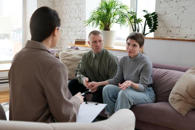 A couple sits on a sofa, engaged in conversation with a person holding a clipboard. The room has a potted plant and bright soft furnishings.