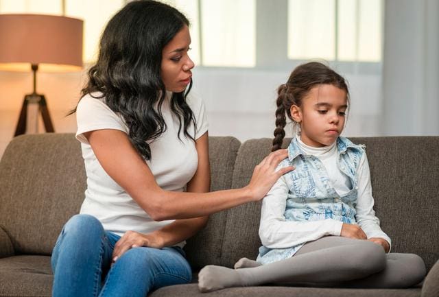 Woman comforts a sad girl sitting on a couch in a living room.