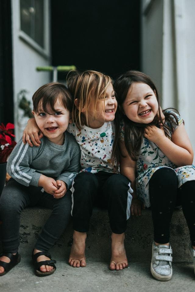 Three children sitting on a concrete step outside, smiling and laughing together.