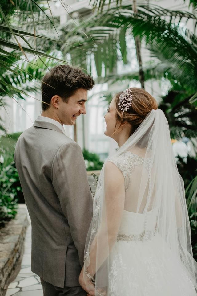 A bride and groom smile at each other in a greenhouse, surrounded by lush green plants. The bride wears a white dress and veil, while the groom is in a gray suit.