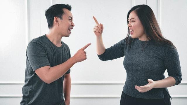 Two people in gray shirts are facing each other, appearing to argue, while pointing fingers in a white-walled room.