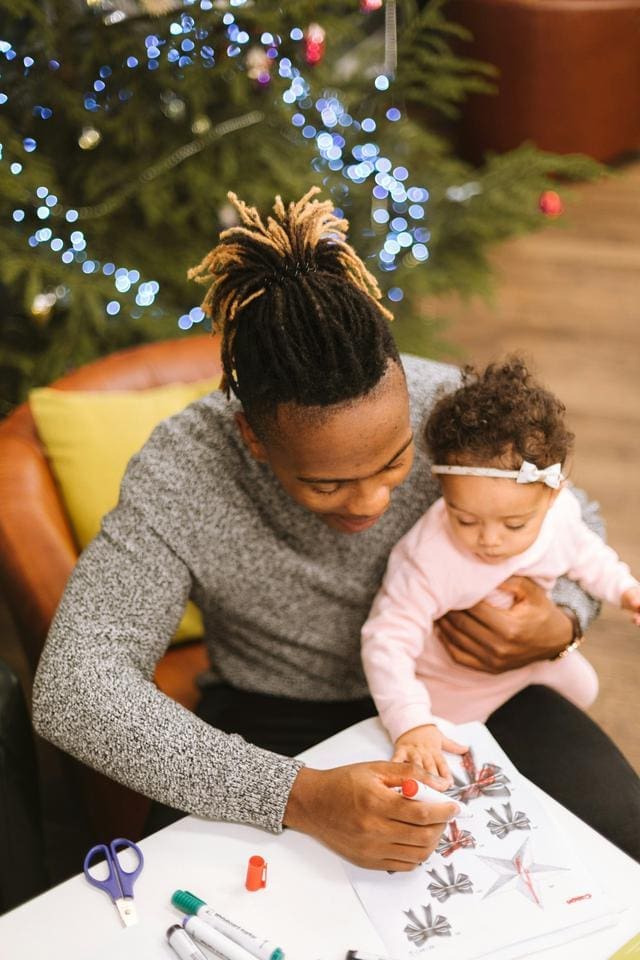 A person and a child color together at a table, with a decorated Christmas tree in the background.