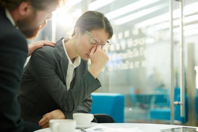 A man comforts a woman in business attire who is sitting and holding her glasses, appearing stressed. They are in a modern office setting with coffee cups on the table.