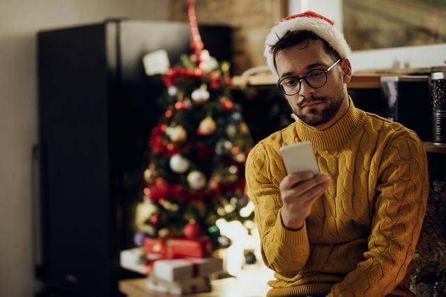 A man in a Santa hat and yellow sweater looks at his phone, with a decorated Christmas tree and gifts in the background.