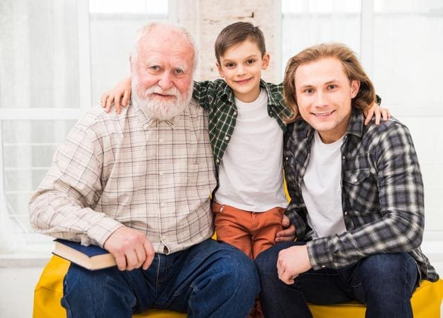 Three generations of a family pose together, with an elderly man holding a book, a young boy in the center, and a middle-aged man on the right, all smiling.