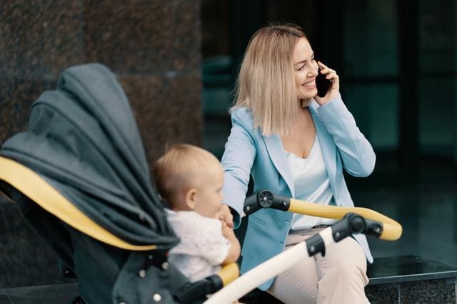 A woman in a blue blazer talks on her phone while sitting next to a baby in a stroller.