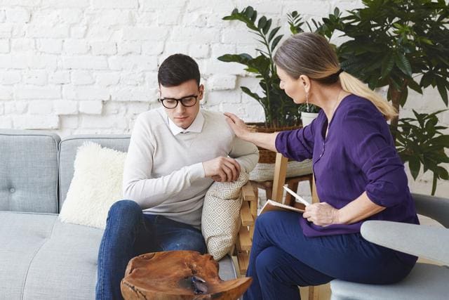 A woman in a purple blouse comforts a man in glasses sitting on a sofa.