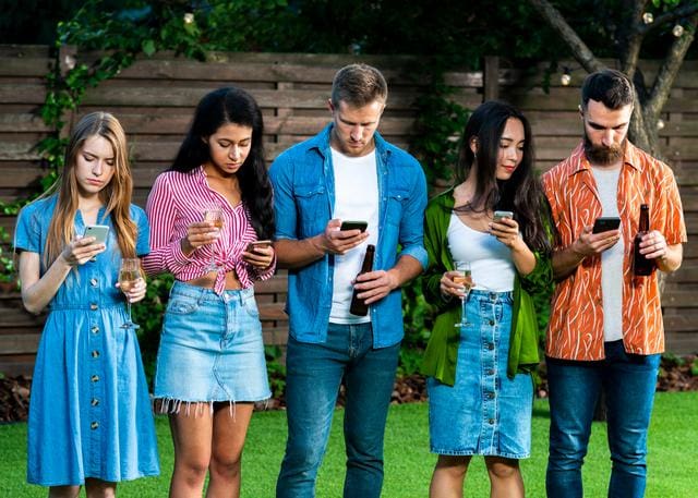 Five people stand in a line outdoors, each looking at their phones. They are holding drinks and are dressed casually. A wooden fence is in the background.