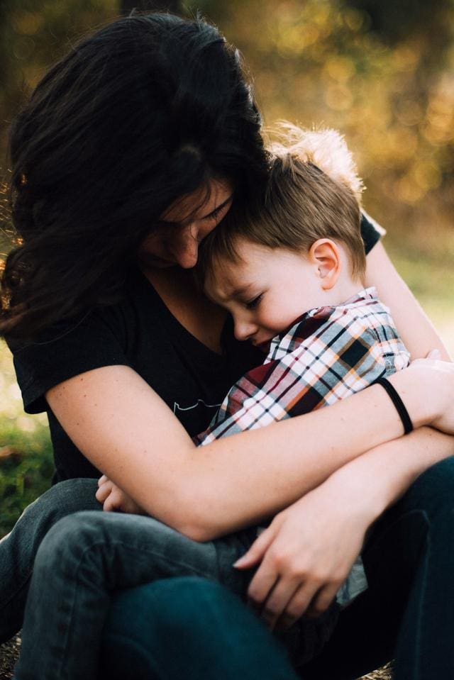 A woman hugs a child outdoors. The child is wearing a plaid shirt and has eyes closed, resting against her. The background is blurred with warm tones.