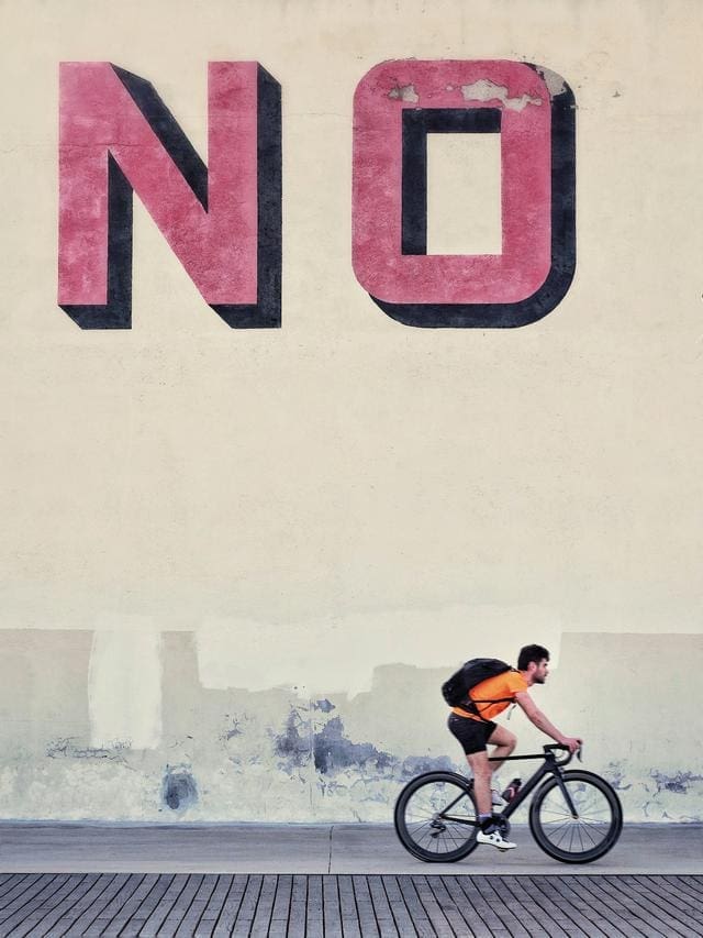 A cyclist rides by a large wall with the word "NO" painted in red above him.