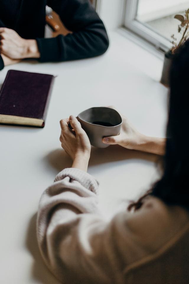 Two people seated at a table with a book and a cup. One person holds the cup with both hands.