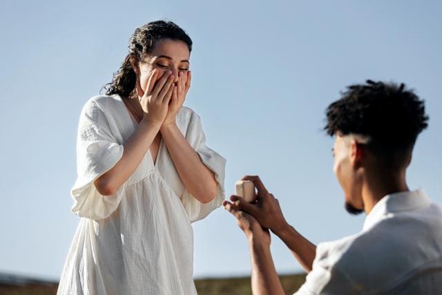 A person kneels, holding an open ring box, proposing to a surprised individual who covers their mouth with their hands. They are outdoors on a clear day.