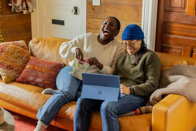 Two people sitting on a couch, laughing. One is holding a gift bag, the other is using a laptop. Colorful pillows are on the couch.