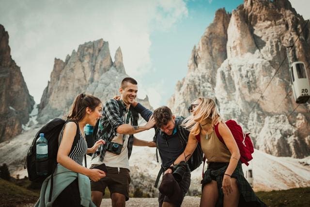 A group of five people with backpacks are laughing and interacting in a mountainous area with rocky peaks in the background.