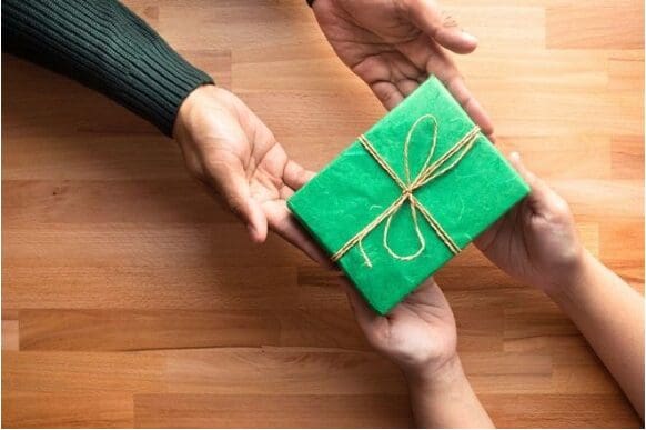 Two hands gift a small, green, wrapped present with twine to another pair of hands over a wooden surface.