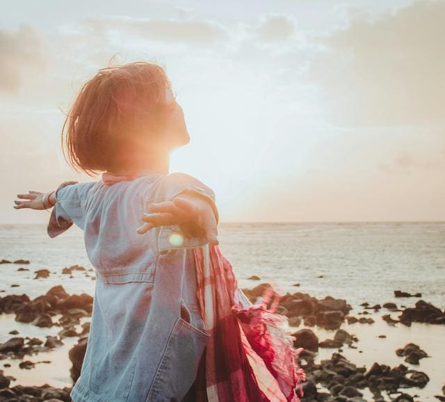 A person with short hair stands on a rocky beach, arms outstretched, facing the sunset. They wear a light blue jacket and hold a red scarf, with the ocean in the background.