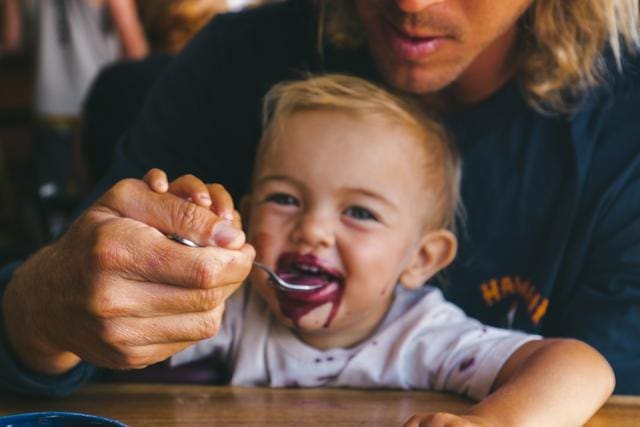 An adult helps a smiling toddler eat with a spoon, as the toddler has purple-colored food around their mouth.