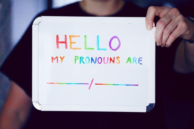 Person holding a whiteboard with "Hello, my pronouns are" in colorful letters, followed by blank lines.