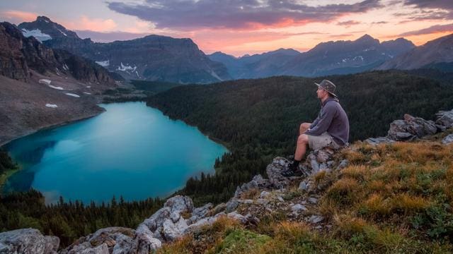 Person sitting on a rocky cliff overlooking a turquoise lake and forested mountains at sunset.