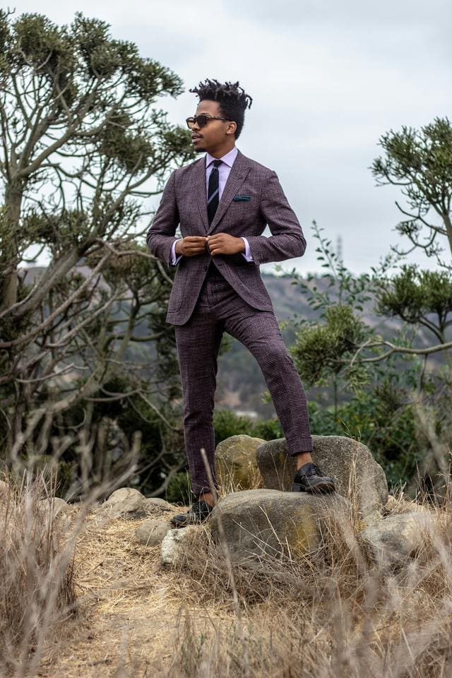 Man in a suit stands on rocks in a natural outdoor setting with trees and hills in the background.