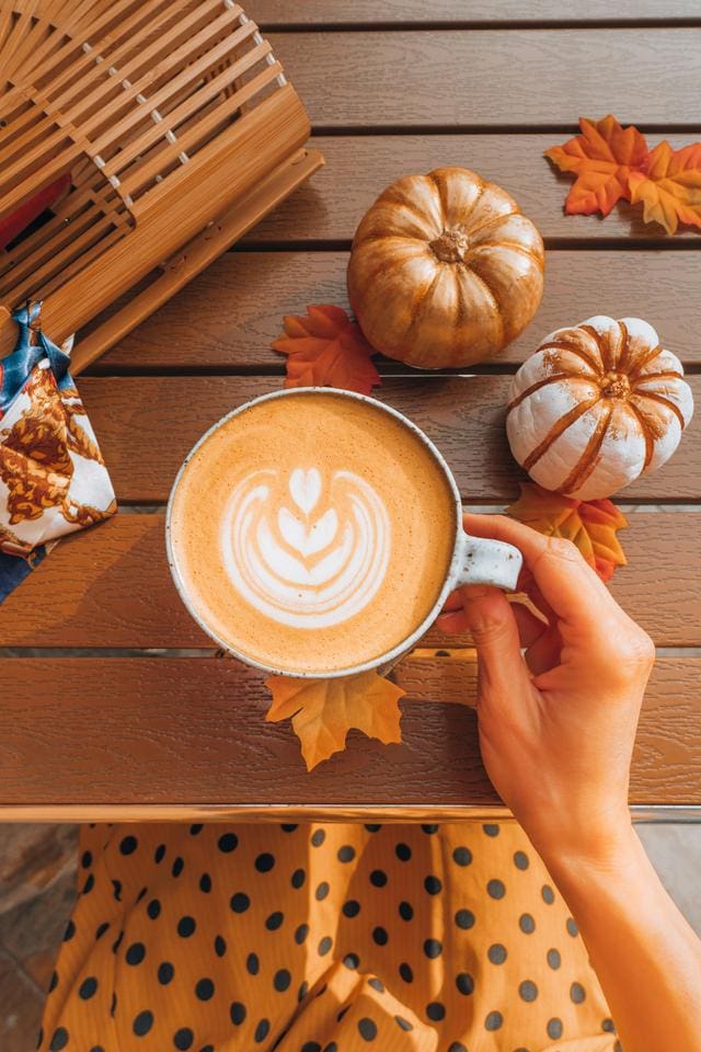 A hand holds a latte with frothy art on a wooden table, surrounded by small pumpkins, autumn leaves, and a snack bar.