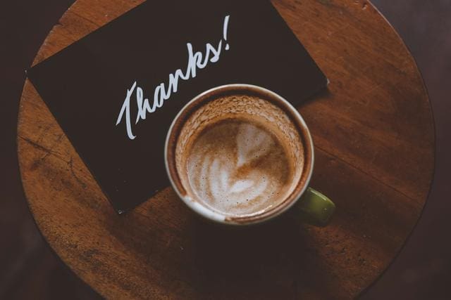 A latte with light froth is on a wooden table next to a black card with "Thanks!" written in white script.