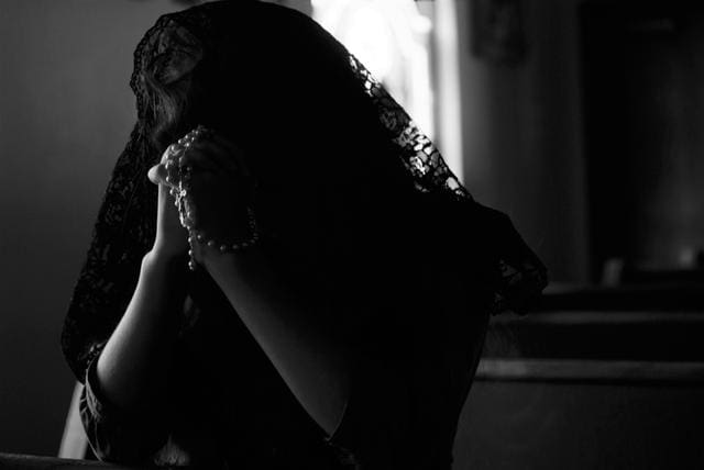 A person wearing a dark lace veil holds a rosary, seated in a dimly lit room.