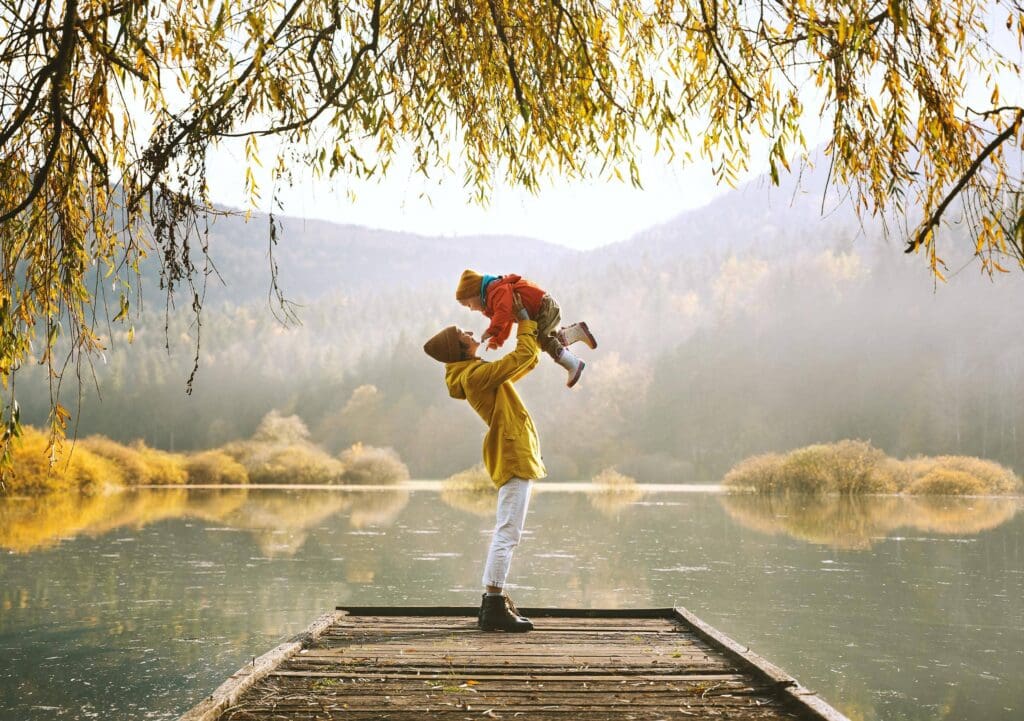 A person lifts a child on a wooden dock by a serene lake, surrounded by autumn trees and misty mountains.