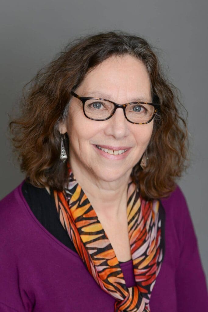 A woman with curly brown hair and glasses smiles, wearing a purple top and a colorful scarf, against a gray background.