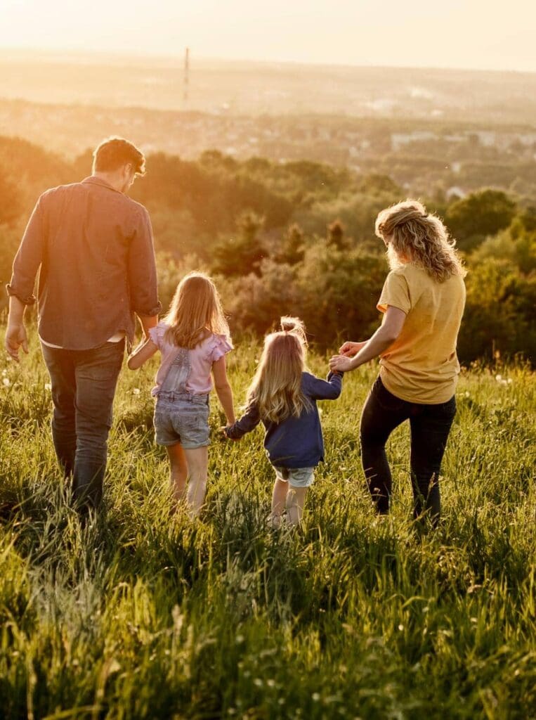 A family of four walks hand in hand through a grassy field at sunset, with a scenic landscape in the background.