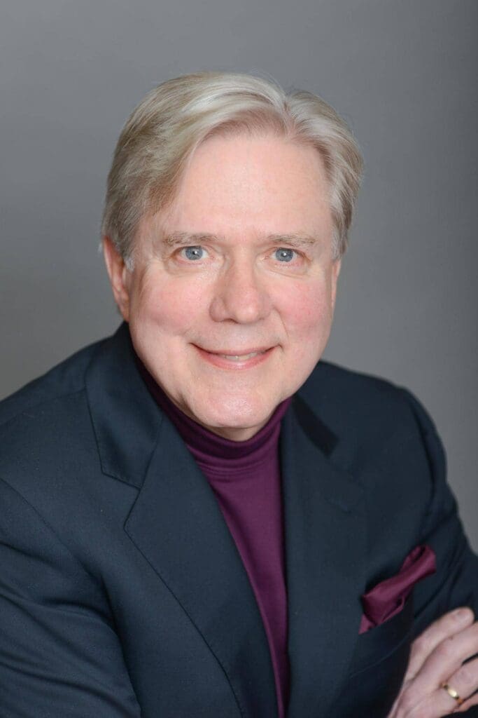 A person with light hair wearing a dark suit and maroon turtleneck smiles at the camera.