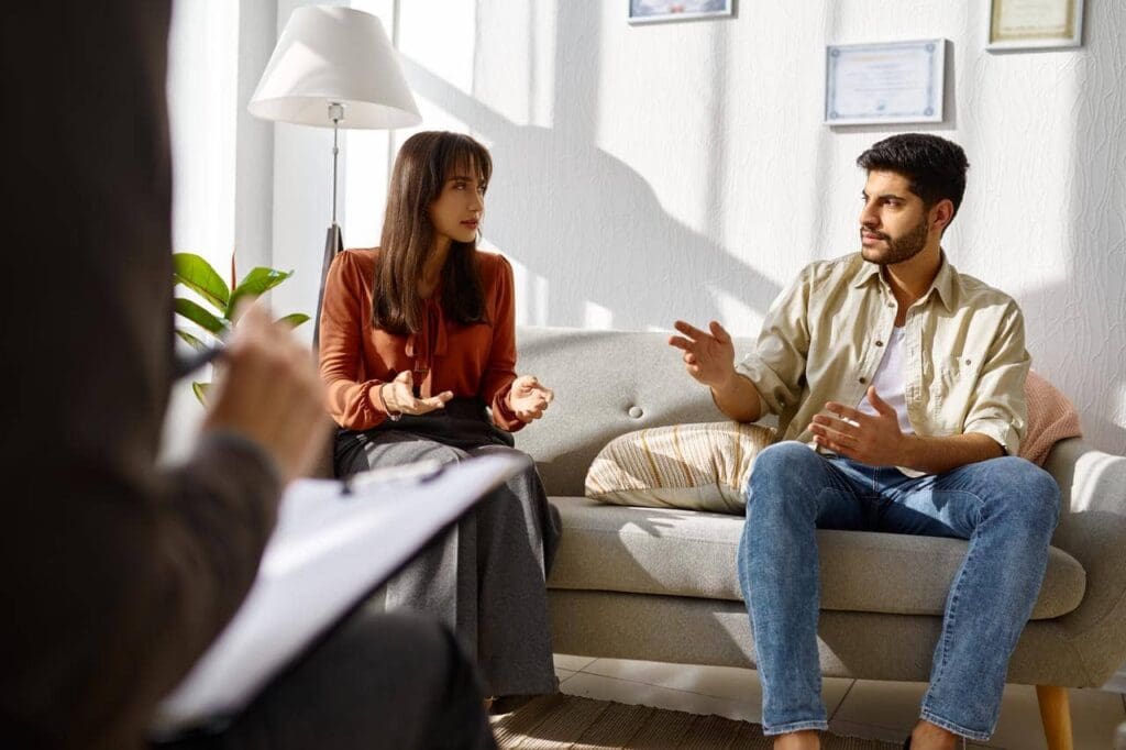 Two people sit on a sofa having a discussion, gesturing with their hands. A third person in the foreground holds a clipboard, observing.