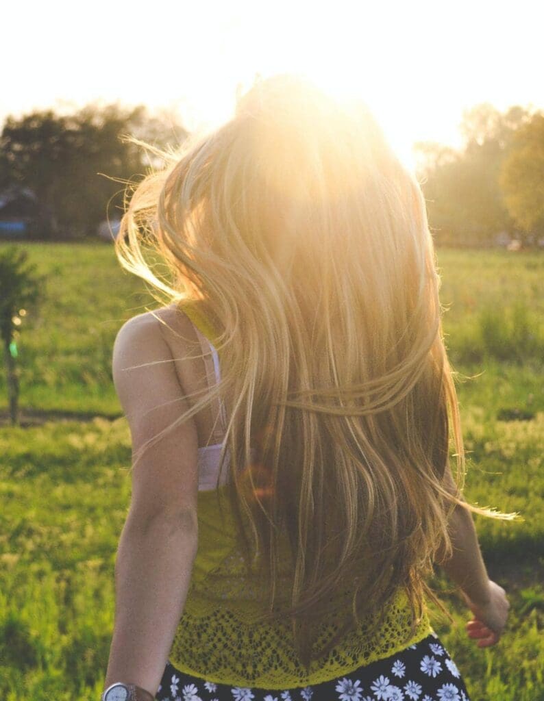 Person with long blonde hair stands in a sunny field, facing away.