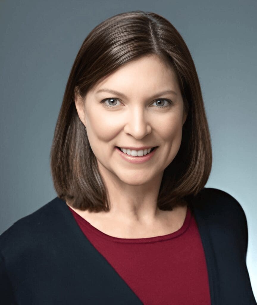 Woman with shoulder-length brown hair wearing a black and red top, smiling against a grey background.