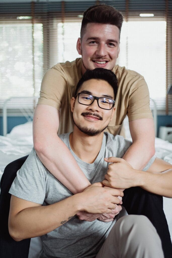 Two people sitting on a bed. The person in the front is wearing glasses and a gray shirt, while the person behind has a beige shirt and is embracing the front person.