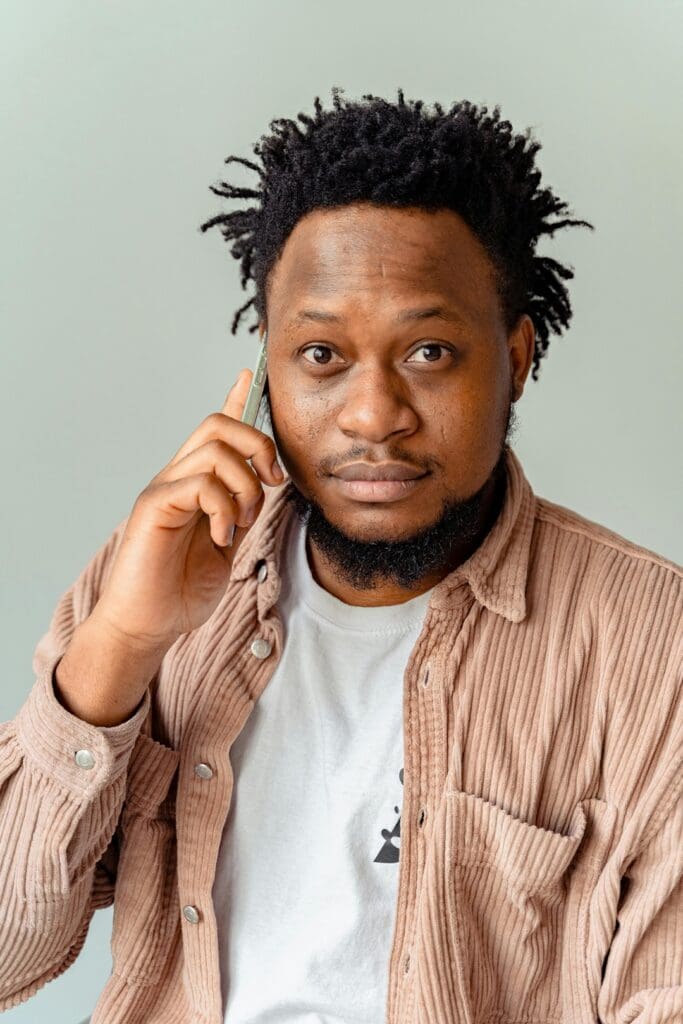 Person with short, curly hair and a beard is holding a phone to their ear, wearing a white t-shirt and a beige corduroy shirt.