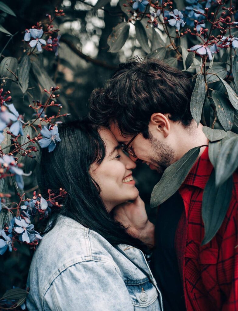 A couple stands closely, foreheads touching, surrounded by lush greenery and purple flowers.