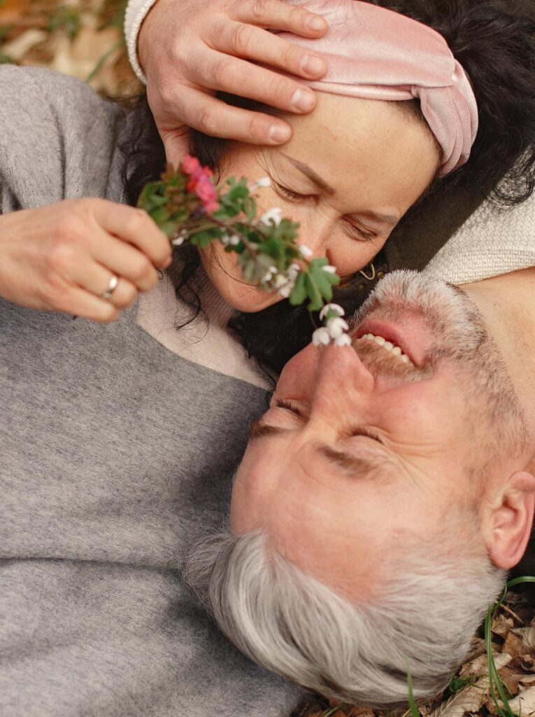 An older couple lies on a blanket outside, smiling and holding flowers. The man has gray hair and a beard, and the woman wears a headband.