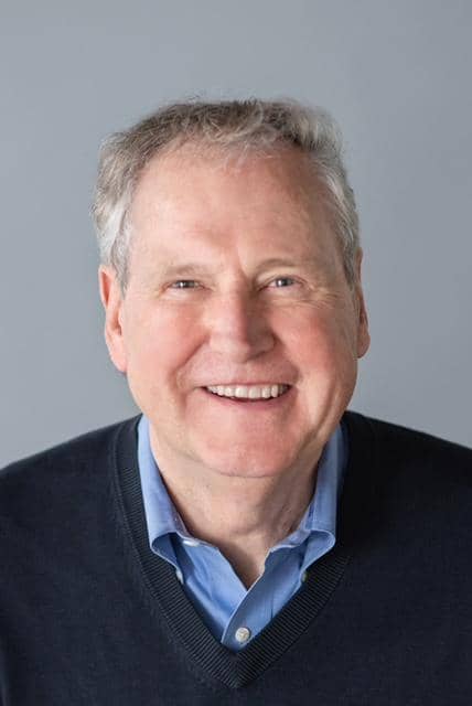 Portrait of a smiling man with short gray hair, wearing a blue shirt and dark sweater, against a gray background.