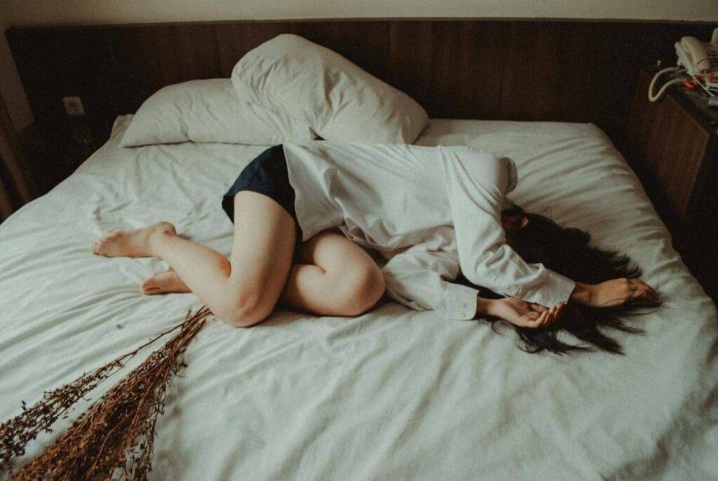 Person lying on a bed with face turned away, wearing a white shirt and dark shorts. Dried flowers are placed on the bed beside them.