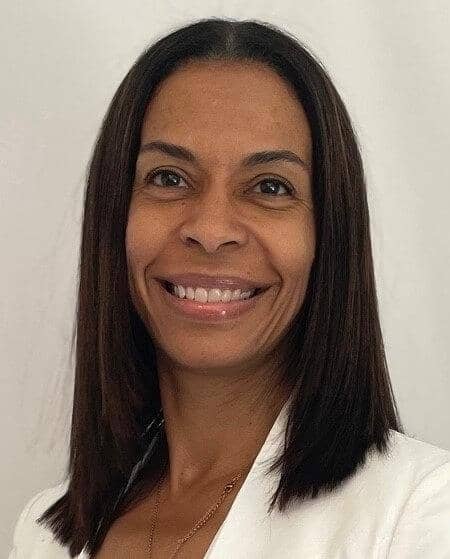 A woman with straight brown hair smiles at the camera, wearing a white top.