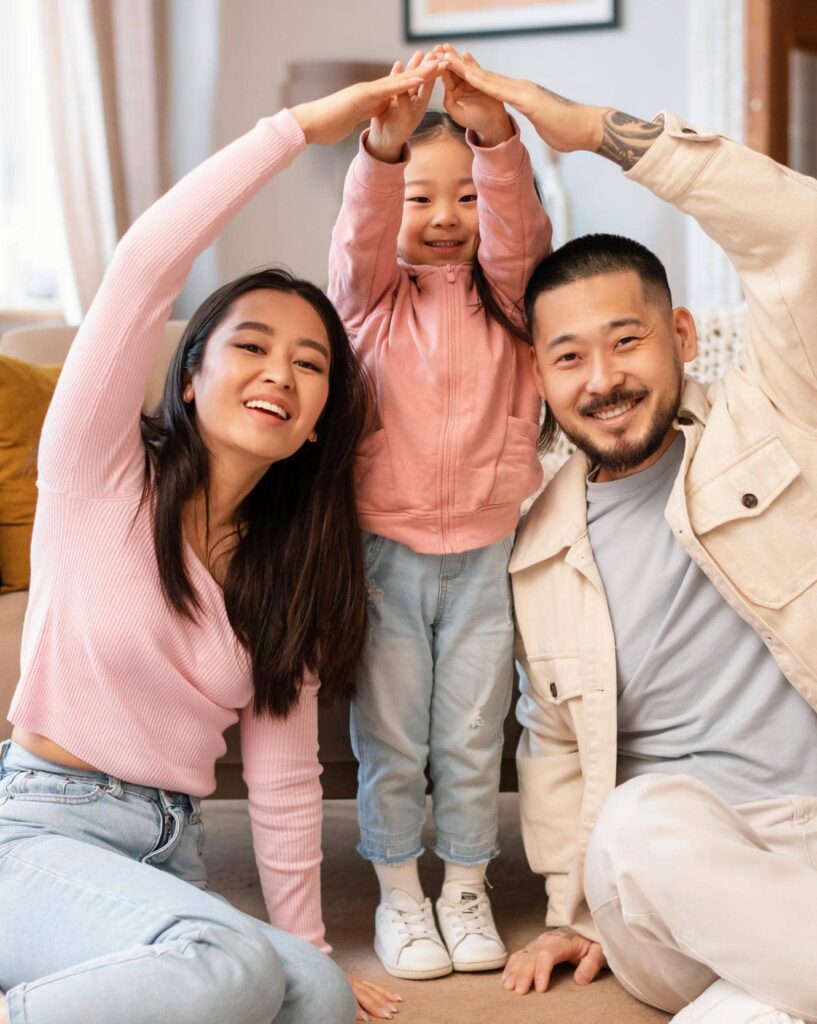 A family of three poses indoors, forming an arch with their arms. The child is in the middle, smiling, dressed in a pink hoodie and jeans. The parents sit on either side, also smiling.