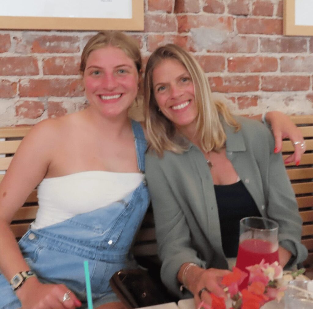 Two women sitting on a bench against a brick wall, smiling. One is wearing a white top with denim overalls, and the other a green shirt. A pink drink is on the table in front of them.