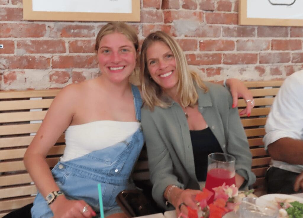 Two people sitting on a bench in a restaurant, smiling at the camera. A brick wall is in the background, and there are drinks and a flower arrangement on the table.