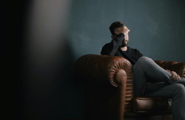 A person sits on a brown leather couch, resting their head on one hand against a dark green wall.