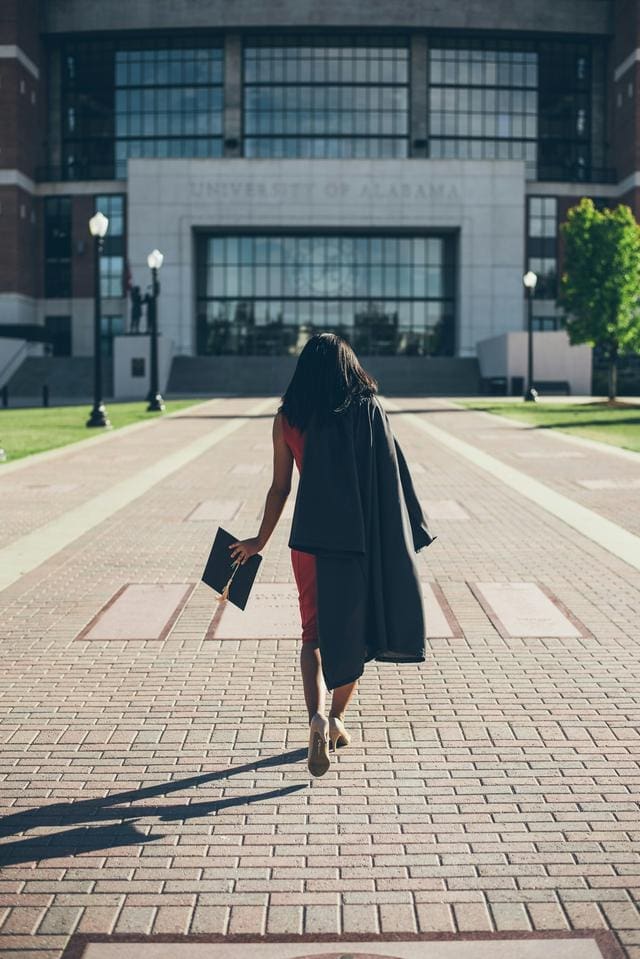 Person walking toward a university building, holding a graduation cap and gown.