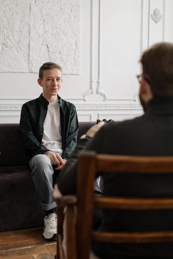 A young person sits on a couch, facing an adult seated in the foreground. They are in a room with white walls and wooden flooring.