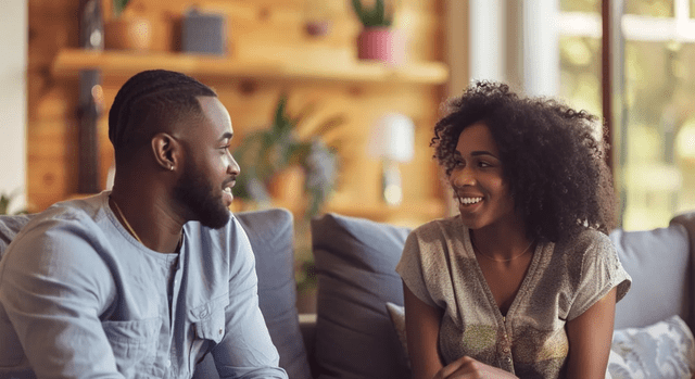 Two people sit on a sofa, smiling at each other in a cozy, well-lit room with plants and wooden shelves in the background.