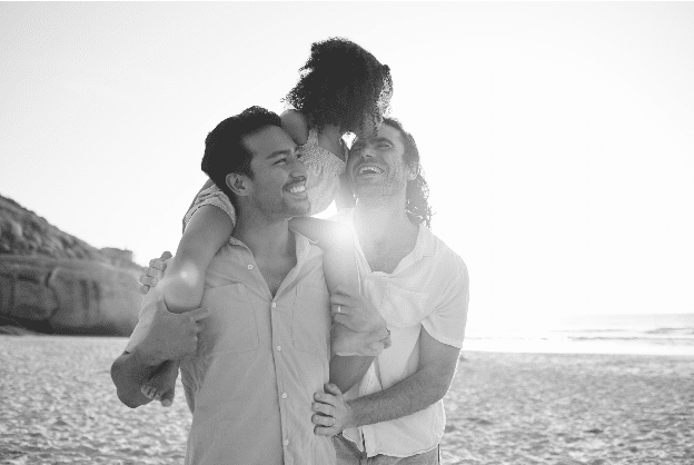 A child sits on an adult's shoulders, with another adult beside them, all smiling on a sunny beach.