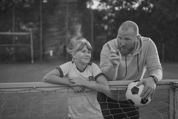 Coach and young soccer player lean on a goalpost. Coach gestures while holding a soccer ball. They are having a conversation on the field.